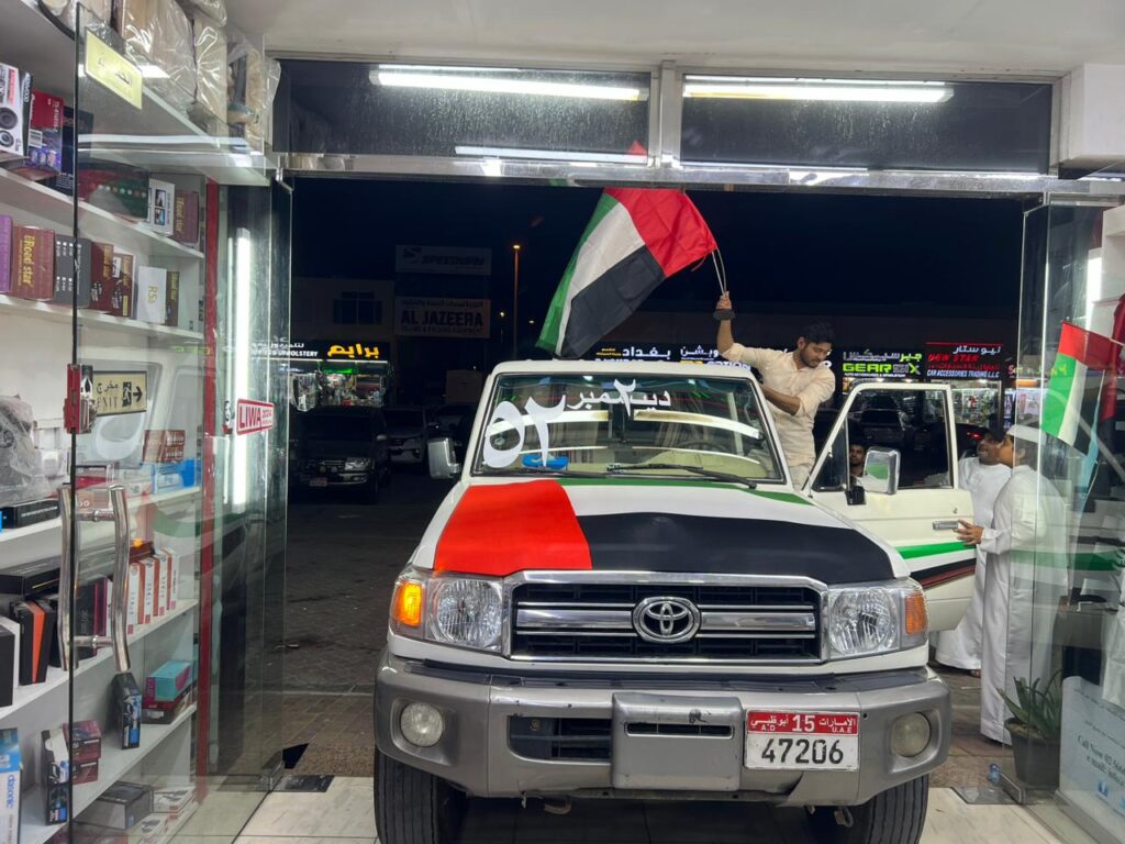 A man stands with the UAE flag in front of AlBadiya car shop, symbolizing patriotism and community engagement.