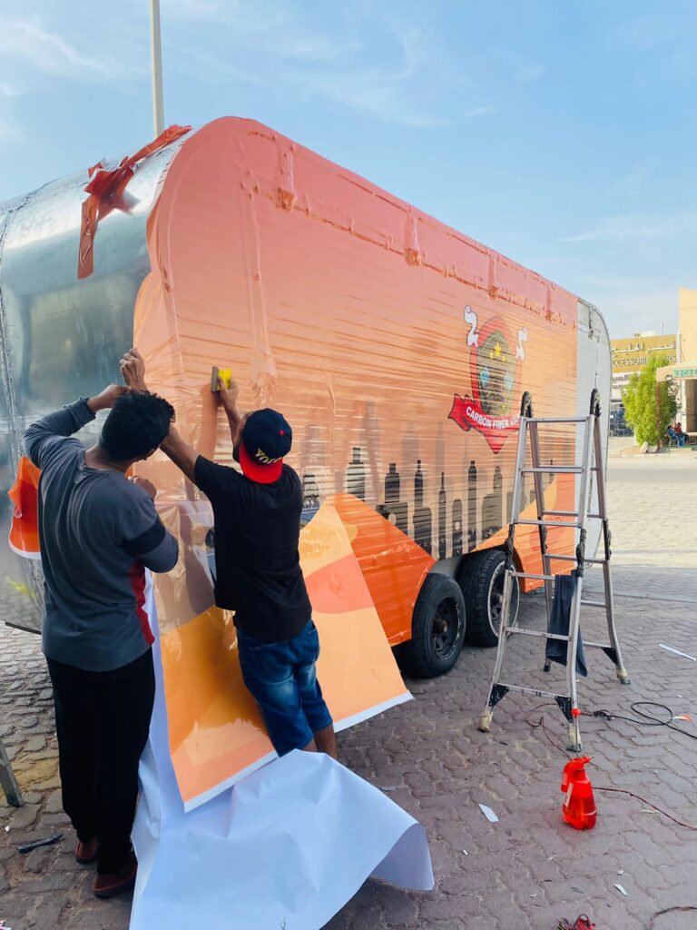 Two men applying a vibrant orange and black car wrap to a trailer, showcasing their painting skills and teamwork.