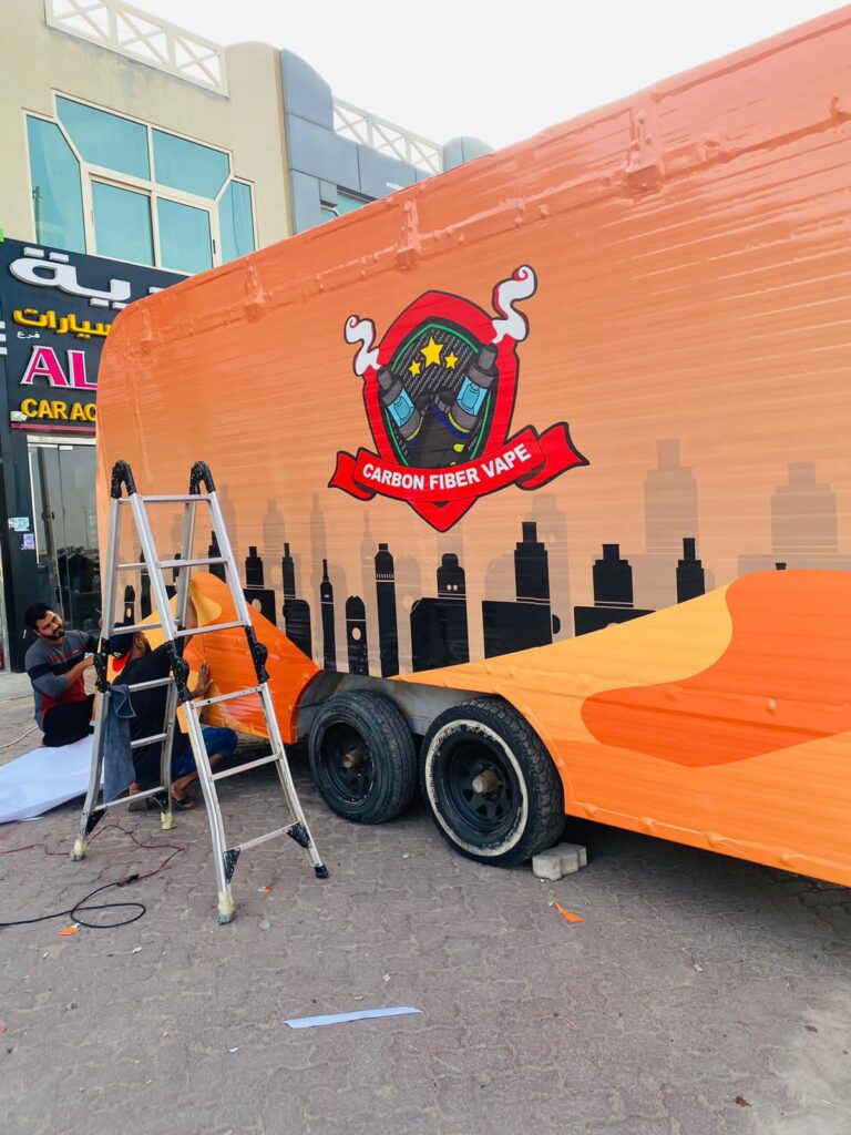 Two men apply a car wrap to a large orange trailer, focusing intently on the intricate details of the design.