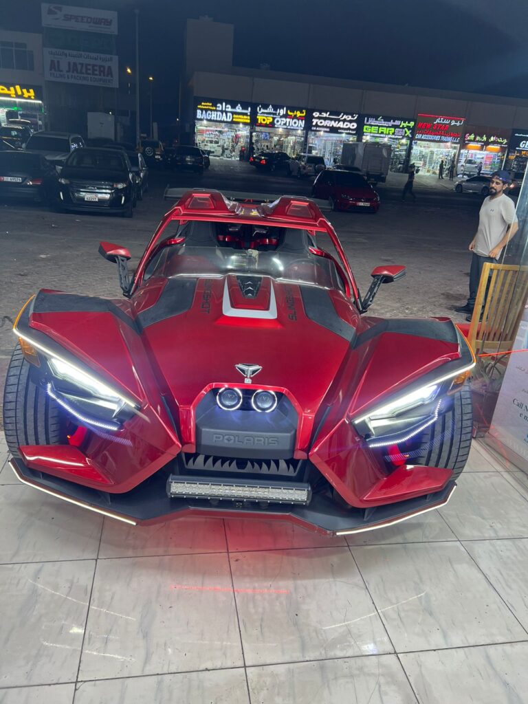 A vibrant red sports car featuring a unique wrap is parked in front of the shop AlBadiya, highlighting its sporty appeal.