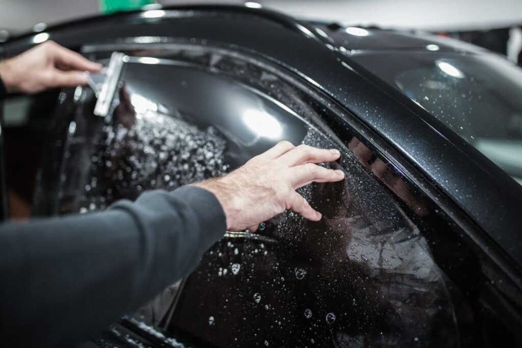 A man is washing the tinted window of a black car, enhancing visibility and preserving the car's sleek look.